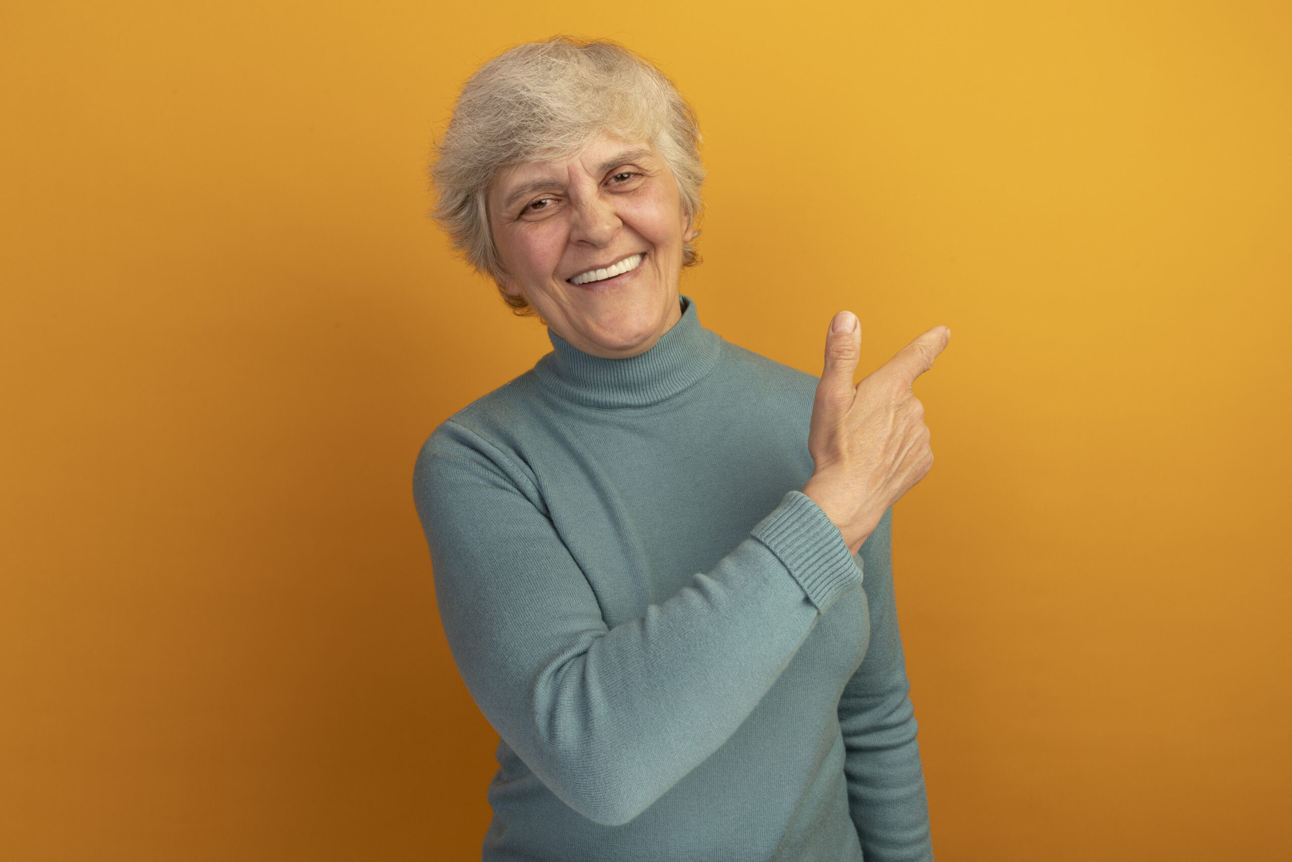 joyful old woman wearing blue turtleneck sweater looking at camera pointing at side isolated on orange background with copy space